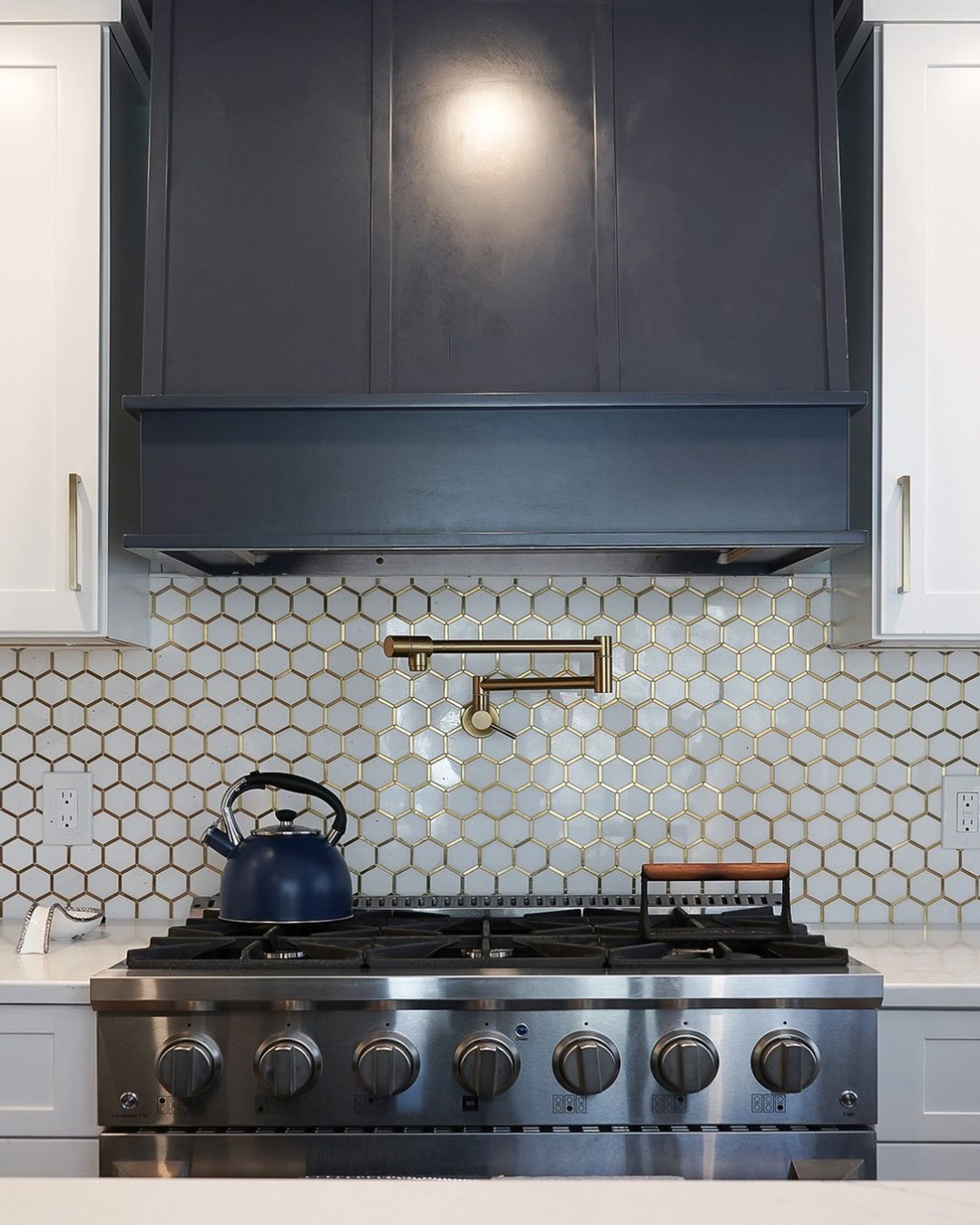 Close-up of modern kitchen remodel featuring stainless steel gas range, navy blue custom range hood, gold pot filler faucet, and white hexagonal tile backsplash with gold accents — Central Connecticut Building and Remodeling, Berlin, CT.