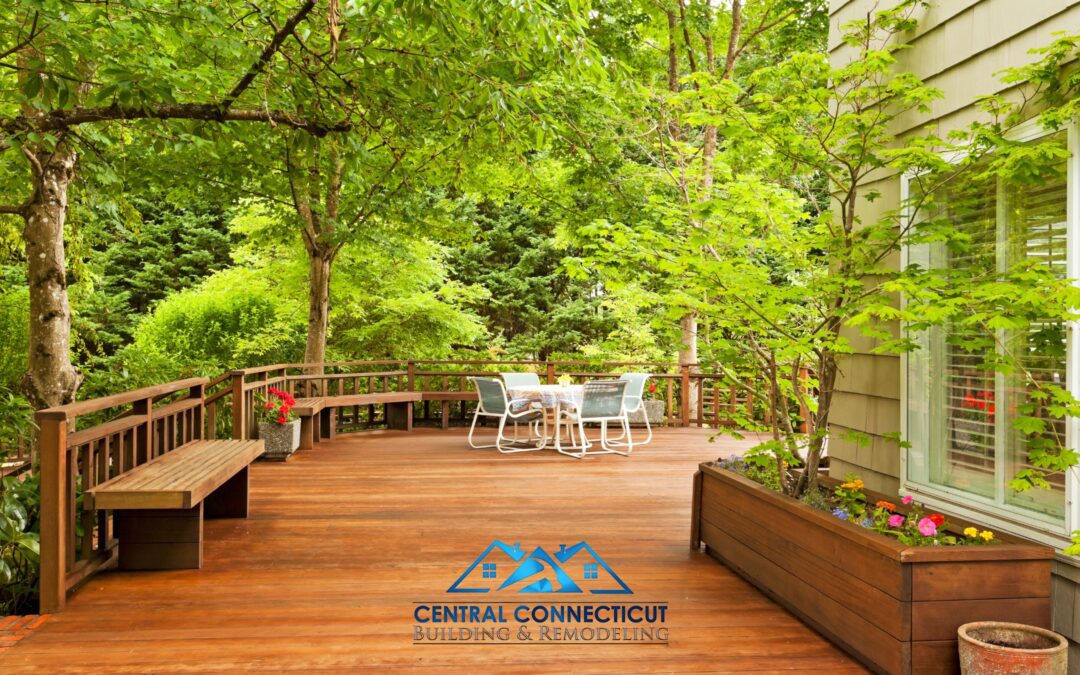 Finished custom wooden deck surrounded by lush green trees, featuring built-in benches, planter boxes with flowers, and a table with chairs — designed and built by Central Connecticut Building & Remodeling in Berlin, CT.